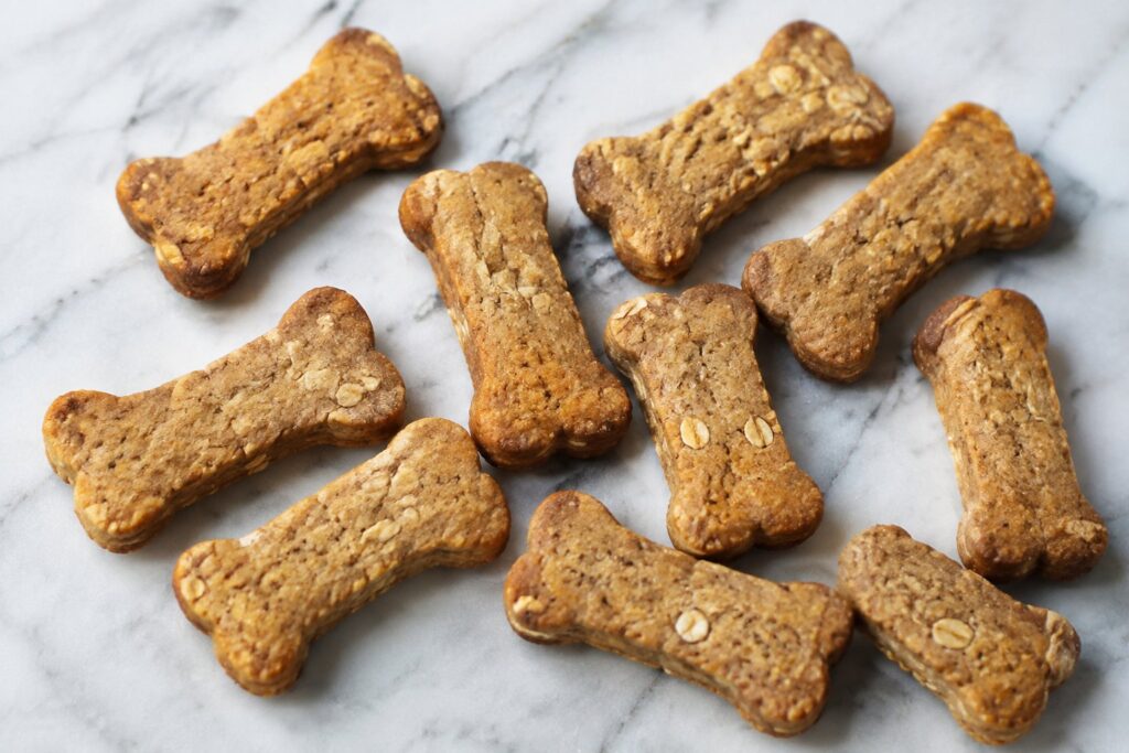 Golden-brown bone-shaped peanut butter applesauce dog biscuits with visible oat flakes scattered on white marble.